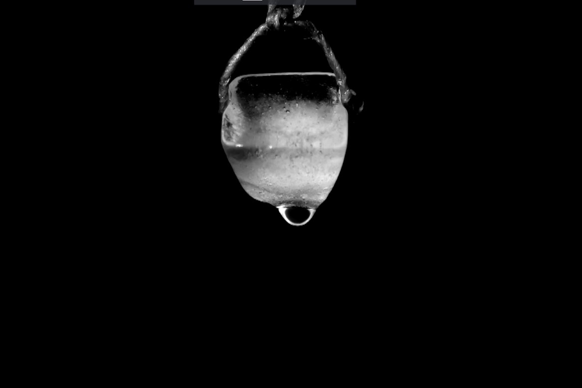 A black and white image of an ice cube hanging from the ceiling with a droplet of water about to fall off against a plain black background