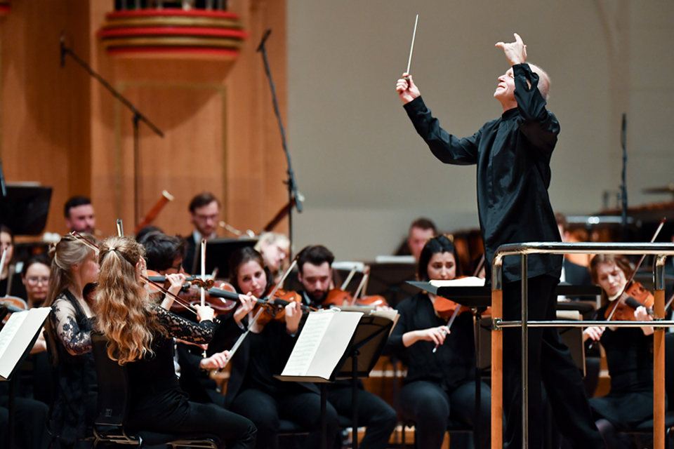 A male conductor, conducting an orchestra, with multiple students performing on string, brass and woodwind instruments. 