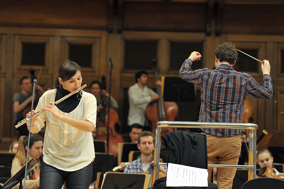 A person playing the flute in front of an orchestra, with a conductor
