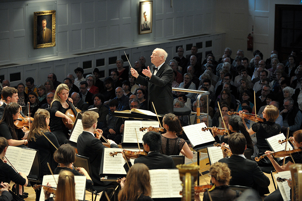 A older man with white hair, conducting in the conductors pit, with students performing in front of him, and the audience behind him.